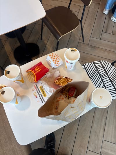 This image presents a top-down view of a white table laden with remnants of a McDonald's meal, set within an indoor restaurant in San Buenaventura, Mexico. The floor features light wood-patterned tiles arranged in a herringbone pattern, suggesting a modern fast-food dining environment.

On the table, several McDonald's items are visible. These include multiple white beverage cups, mostly consumed, some with yellow accents. There are two portions of french fries, one in a classic red McDonald's carton with the "W" logo and another in a larger paper bag that also contains other food packaging. A red sauce packet, partially obscured but showing "Salsa B," and a ketchup packet are present. An empty "Chicken McNuggets" box, labeled "10," lies on the table, indicating a 10-piece order. A large, brown paper bag, likely the main takeout bag, is crumpled and contains additional packaging. Discarded paper napkins are also scattered on the table.

Partial views of two individuals are visible. At the top-left, a person's legs are seen, wearing blue jeans and white sandals. At the bottom-right, a person's dark pants and black shoes with a silver buckle are visible. The chairs around the table include one with a distinctive black and white geometric pattern on its backrest and another with a dark brown wooden seat and black metal frame.

A notable detail on the table is a circular white placemat or coaster featuring Spanish text related to a loyalty program: "MiM," "Regístrate y suma puntos para canjear por beneficios" (Register and earn points to redeem for benefits), and "$1=10 puntos." The McDonald's "W" logo is consistently displayed across the packaging. The scene depicts a casual meal at a McDonald's, possibly after consumption, given the partially empty containers and discarded items.