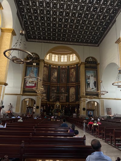 The image captures the interior of a church, likely located in San Salvador, El Salvador. Rows of wooden pews are visible, occupied by individuals attending a service or prayer. At the far end is a large altar with framed paintings and intricate architectural details. Circular chandeliers hang from the high ceiling. The ceiling has an ornate geometric pattern. The atmosphere suggests a place of worship and reverence.