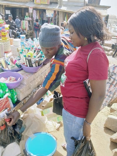 A daytime outdoor market scene is depicted in the city of Kungulu School, Zambia. The foreground features two individuals engaged at a vendor's stall, with general market activity occurring in the background.
On the right side of the frame, a female individual with dark complexion and dark, shoulder-length hair is visible in profile. She wears a red short-sleeved t-shirt with a white and green graphic on the left chest area, light blue denim jeans, a gold-colored stud earring in her right ear, a silver-colored chain strap draped over her right shoulder, and a black beaded bracelet on her left wrist. She looks downwards towards the stall. Her left hand holds a black plastic shopping bag.
On the left, partially obscured by the female, a male individual with dark complexion wears a grey knit beanie and a multi-colored striped long-sleeved shirt featuring blue, orange, green, and brown stripes. A gold-colored watch is visible on his left wrist. He is leaning forward over the stall, with his right hand extended downwards, appearing to manipulate contents within a black plastic bag situated among other items on the ground or lower stall area. His left arm is bent at the elbow, resting near the merchandise.
The vendor's stall, a simple structure, displays numerous small packaged goods and containers. Prominent items include multiple plastic bowls; one purple bowl contains an assortment of white-capped and black-capped cylindrical objects, consistent with pens or small bottles. Another purple bowl holds small, colorful items. Stacks of rectangular packaged goods and small green packages are arranged on and around the stall surface.
The ground directly below the stall and subjects is unpaved, appearing dusty or sandy and irregular. A light blue plastic bucket containing a light blue liquid is positioned on the ground beneath the stall. Various discarded plastic bags (beige and black) and cardboard boxes are scattered on the ground around the transaction area.
In the background, indistinct market stalls with corrugated metal roofs and visible merchandise line what appears to be a street or pathway. Other individuals, including one on a motorcycle to the far left, are present. Distant vehicles and structures are discernible, indicative of a busy urban or semi-urban thoroughfare. The scene is illuminated by bright natural light.