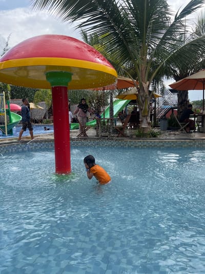 The image displays an outdoor swimming pool facility located in Juwana, Indonesia. A central feature in the foreground is a large, red and yellow mushroom-shaped water fountain actively spraying water into a light blue tiled pool. A young child, wearing an orange shirt and white shorts, stands submerged waist-deep in the pool directly beneath the water feature, with both hands covering their ears or face amidst the splashing water. To the left, an adult male in a dark blue shirt and shorts walks along the poolside. An adult female, wearing a light-colored top and dark hijab, stands further back near the water feature. In the background, several large palm trees rise above orange and yellow sun umbrellas, beneath which multiple individuals are seated at wooden tables. Visible structures include parts of green water slides and a white staircase leading to an upper platform. The sky is overcast.