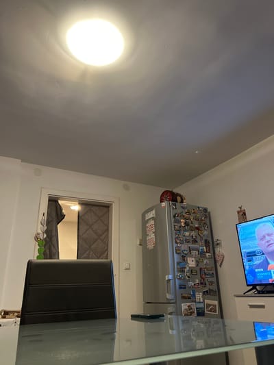 This image depicts an indoor scene, specifically a residential area, located in Benningen, Germany. The perspective is from a seated position at a reflective dark-surfaced table, possibly glass, which occupies the lower third of the frame and shows reflections of the ceiling light and television.

The primary light source is a prominent, bright circular ceiling light positioned in the upper center. The ceiling and walls are white. On the upper left wall, above a doorway, two circular, white speaker-like fixtures are mounted.

In the immediate foreground, a dark, possibly black, chair back is visible, facing away from the viewer. A dark-cased smartphone rests horizontally on the table surface to the left of the chair.

To the right, a silver-colored refrigerator stands against the wall, heavily adorned with numerous souvenir magnets and a few paper items, including what appears to be a calendar. A decorative cylindrical object with a star or compass design on its lid, along with another small container, rests on top of the refrigerator.

Further right, a black flat-screen television is displaying what appears to be a news broadcast. The screen shows a close-up of a Caucasian man's face, with light hair, looking towards the right. Partial Cyrillic text ("НОВИНИ," meaning "News") and a vehicle with "POL" visible (suggesting police) are also displayed on the screen. The television is supported by a white, low-profile cabinet.

To the left, a doorway leads into an adjacent room, partially obscured by a dark grey, quilted curtain or divider. A light source is visible within the room beyond the doorway. A decorative hanging item composed of multiple grey heart-shaped elements is suspended near the doorway.

No individuals are actively present or interacting within the immediate captured space; the visible person is an image on the television screen.