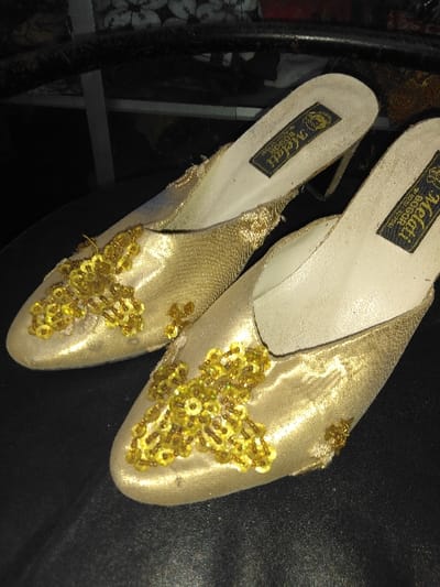 This image depicts a pair of women's gold-colored high-heeled mules resting on a dark, textured surface within an indoor setting in Kruibeke, Belgium. Each shoe features an off-white insole marked with a black rectangular label displaying the text "Melati BOGOR" and a phone number "0251-321155." The uppers are crafted from a shimmering gold fabric, possibly satin, and are embellished with clusters of gold sequins and beads arranged in distinct floral or abstract patterns on the vamp. The insoles show some particulate matter or dust. The background is out of focus, revealing a dark environment with a glimpse of a patterned textile object in the upper right and a light-colored vertical support structure towards the center. No individuals, actions, or direct interactions are visible.