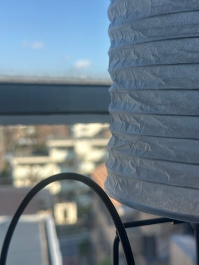 The image captures an indoor perspective looking out onto an urban landscape in Nakano, Japan. In the foreground on the right, a significant portion of a white, pleated paper lantern or lamp shade is visible, characterized by its textured, horizontally folded surface. A slender, black metal support structure is partially seen beneath the lantern. To the bottom-center and left, a curved, black metal arch partially obstructs the view of the outdoor environment.

The background displays a blurred urban scene under a clear blue sky dotted with minimal white clouds. A prominent, dark, horizontal structure, consistent with an elevated railway line or bridge, traverses the upper midground. Below this elevated feature, numerous multi-story buildings with light-toned facades are visible, indicative of residential or commercial architecture common in a Japanese city. The scene is devoid of discernible individuals, actions, or specific interactions, illuminated by bright daylight.