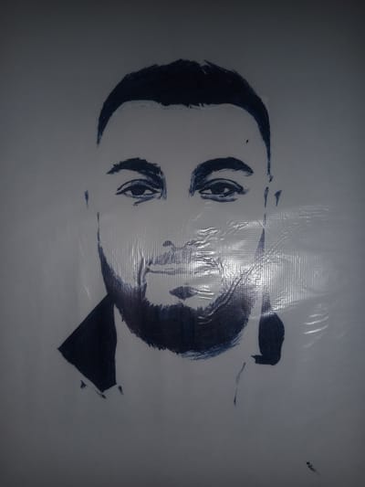 This is a portrait of a man's face, rendered in dark blue ink on a light gray background. The man has dark hair styled with a sweeping fringe, bushy eyebrows, and a beard. His eyes are looking directly at the viewer. The portrait is characterized by a loose, sketch-like style, with visible brushstrokes and an unfinished quality. The image appears to be on a textured surface, possibly a banner or fabric, as indicated by the grid-like pattern and reflections of light, suggesting it is displayed outdoors. The style of the portrait suggests it could be a mural or a large-scale artwork. There is no indication of a specific location in Gulbag, Uzbekistan within the image itself.