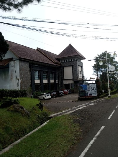 The image displays an exterior daytime scene featuring a multi-story building, an adjacent parking area, and a paved road under an overcast sky. The provided location for this scene is Phoxay, Laos.

The building exhibits a facade primarily composed of dark, textured stone or stone-like cladding, topped with a complex brown tiled roof. A prominent square tower element, also stone-clad and featuring a pyramidal tiled roof, is positioned on the right side of the building, containing a large, recessed rectangular opening. The building's left section presents a lighter-colored wall with visible staining or weathering. Multiple large windows with dark frames are present on the building's visible face.

In front of the building, an asphalt parking lot contains several vehicles: a white hatchback, a black SUV, and multiple dark red or maroon vehicles. Near the entrance to this lot, a large rectangular sign displays an image of an interior space and text that includes "350 ribu". Adjacent to this sign, a white pole supports multiple ornate lamppost lights and an oval sign featuring Arabic script alongside the English word "AL KHAIRA".

The immediate foreground shows a paved asphalt road marked with faded white dashed and solid lines. A narrow strip, possibly a drainage grate or textured surface, runs along the road's edge, separating it from a sloping, green grassy embankment. Several dark rocks are embedded within this grass. Overhead, numerous black power lines or communication cables crisscross the gray sky. Portions of a pine-like tree are visible on the far left and near the lamppost on the right. No individuals are present in the depicted scene.