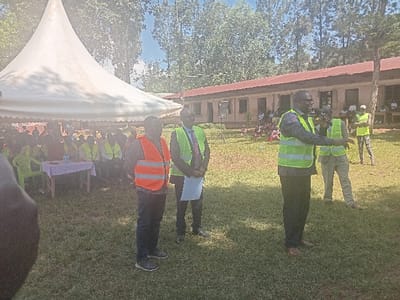 The image depicts an outdoor gathering in Idakho South ward, Kenya, on a sunny day. In the foreground, a vibrant green lawn spans the lower portion of the frame. Three adult males stand prominently in the midground. The leftmost visible male wears an orange reflective safety vest over a dark long-sleeved shirt and dark trousers. Adjacent to him, a male in a bright green reflective safety vest over a dark suit holds white papers. To their right, a third male, also in a bright green reflective safety vest over a dark shirt and dark trousers, is actively speaking into a black microphone held in his right hand while gesturing with his left. A fourth male in a bright green vest and light trousers stands partially visible to the speaker's right.
On the left side of the scene, a large white canopy tent is erected, under which numerous individuals are seated on chairs, many wearing bright green or yellow reflective safety vests. A table covered with a light-colored cloth, holding water bottles, is visible beneath the tent. In the background, a single-story building with a red corrugated iron roof and multiple rectangular windows is present. To the left of this building, a black speaker is mounted on a tripod stand. Beyond the building and the tent, a dense line of tall trees is visible under a clear, bright sky. Several additional individuals, some in bright vests, are visible standing near the background building.