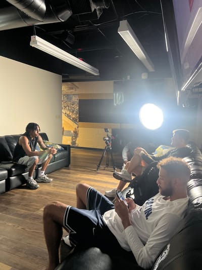 In North Charleston, United States of America, a group of individuals are gathered in what appears to be a studio or media room, likely for a recording or interview. The room features a black leather sofa and a large, dark sectional couch. A man with braided hair, wearing a black tank top and grey shorts, sits on the left side of the sofa, looking to his left. To his right, on the sectional, a young man in a black long-sleeved shirt is seated. In front of him, another man is on his phone, wearing a white long-sleeved shirt with blue lettering and navy shorts. A ring light illuminates the scene from the background, positioned behind a camera on a tripod. The room's walls are a mix of white and dark paneling, with a mural of a basketball court and spectators visible on the back wall. The ceiling is black, with exposed ductwork and fluorescent lighting fixtures. The floor is made of wood-look laminate.