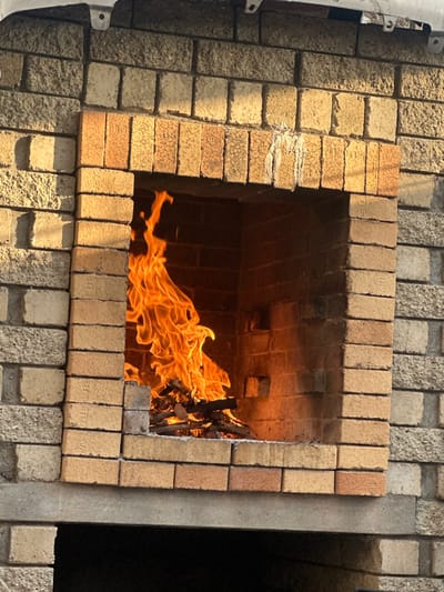 A forensic description of an outdoor brick braai (barbecue) structure, located in Mitchells Plain, South Africa, shows a fire burning within its main rectangular opening. Vibrant orange and yellow flames consume a pile of wood pieces at the base of the firebox. The structure's exterior is composed of light greyish-brown, textured building bricks, while the interior of the firebox and the surrounding lintel are constructed from reddish-brown, smoother bricks. Visible markings on some bricks include "U.G.", "U.D.", "J.S.", and "90". A white, solidified residue streak is present on the underside of the top lintel, directly above the flames. An unseen overhead structure, possibly metallic, is partially visible at the top edge of the frame. The scene is lit by strong directional sunlight, casting shadows inside the firebox. No persons or direct actions are depicted.