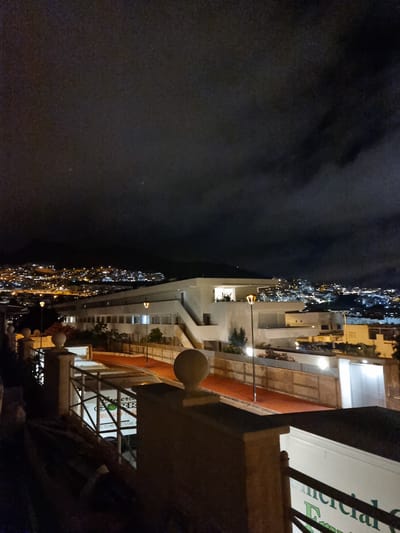 The image depicts a nighttime view of a city, likely Miraverde, Spain, from an elevated position. The foreground features a decorative stone railing and a dark, textured surface. In the mid-ground, a white building with numerous illuminated windows and a staircase is visible, situated on a reddish-orange paved area. Streetlights cast pools of light. Beyond this building, the city sprawls across hills, a dense collection of lights indicating buildings and streets. The sky is dark and overcast with clouds. A white vehicle with green lettering, possibly a delivery truck, is partially visible behind the railing in the foreground. The lettering on the vehicle appears to be "Comercial Frutas y Verduras" and includes telephone numbers.