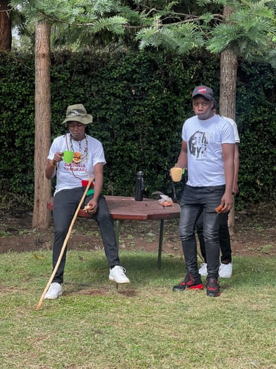 Two men are depicted outdoors in a grassy area with dense green foliage and trees in the background, likely in Makuyu, Kenya. The image is oriented vertically with the subjects standing upright.
On the left, a man stands, looking directly at the camera with a slight smile. He wears a grey cap, a white t-shirt with a black graphic of a person's head and the visible word "MUGWERU," dark jeans, and black and red high-top sneakers. He holds a light-colored cup or container in his right hand.
On the right, a second man is seated on what appears to be a bench or low platform, also looking towards the camera. He wears a light-colored wide-brimmed hat, a white t-shirt printed with "GUCHOKIA RUU MUKARO" and a graphic resembling a sunset or sunrise over mountains. He also wears dark jeans and white sneakers. Around his neck is a lanyard or ribbon in red, green, and black, featuring the text "I AM KENYAN." He holds a green cup in his left hand and a wooden walking stick, which rests diagonally across his body.
Between the two men, a wooden picnic table or bench is visible, with a dark object (possibly a camera or speaker) and some orange items (perhaps food) resting on it. The scene suggests a casual outdoor gathering or break on a bright, sunny day. Both individuals appear relaxed.