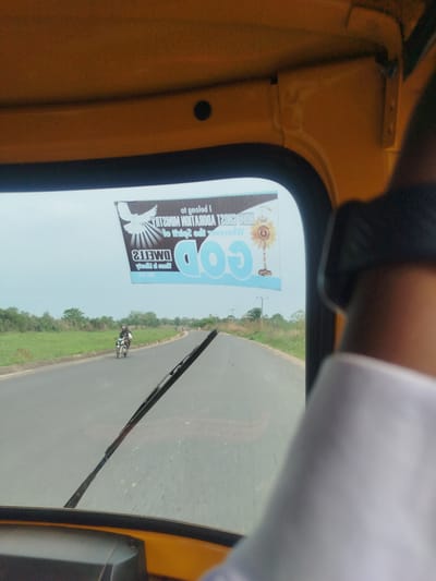 The image displays a first-person perspective from inside a yellow vehicle, likely a tricycle (keke NAPEP), traveling along a paved road in Ukwulu, Nigeria. The vehicle's dashboard and a portion of the steering apparatus are visible on the left, while a person's arm, dressed in a white sleeve, partially obstructs the right side of the view. A single wiper blade rests on the windscreen.
A prominent banner or sticker is affixed to the upper section of the windscreen, appearing in reverse from the viewer's position inside the vehicle. This religious-themed banner features a large "GOD" in blue lettering, a white dove symbol, and a sun-like emblem (possibly a monstrance or sunflower). The reversed text on the banner includes phrases such as "I belong to", "HOLY GHOST ADORATION MINISTRY", "Where the spirit of the Lord", and what is likely "God is able". The reversed word "DMETTE" is also visible.
Beyond the windscreen, the road extends into the distance, bordered by lush green fields and scattered trees under a bright, overcast sky. In the middle ground, a motorcyclist is visible, riding away from the vehicle. The scene captures a daytime journey through a rural or semi-urban landscape, devoid of discernible emotional cues.
