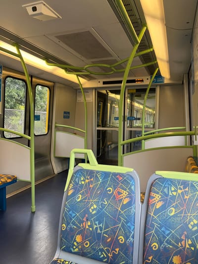 The image displays the interior of an empty train carriage, rotated 90 degrees clockwise. The carriage features rows of seats with a distinctive and colourful patterned upholstery in shades of blue, yellow, orange, and red, adorned with abstract geometric and symbolic designs. The seat frames and numerous overhead and vertical handrails are a bright lime green, providing a prominent accent against the neutral grey and white interior panels of the walls and ceiling.

No people are visible, indicating the carriage is empty. Daylight streams through the rectangular windows, revealing lush green foliage and trees outside, suggesting it is daytime. The floor is covered in a dark, possibly grey or blue, material. Along the ceiling, strip lighting illuminates the interior.

Notable details include a small digital display with amber characters, possibly showing "13" or a time, which is partially visible. Several small blue signs with white pictograms are affixed to the walls, likely indicating information such as priority seating or general instructions, though the text on them is illegible. The design of the carriage, particularly the unique seat pattern and green handrails, is characteristic of metropolitan trains found in Melbourne, Australia, consistent with the provided location context of Northcote.