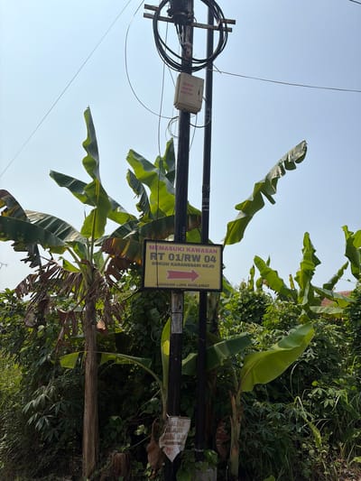 The image depicts a utility pole with an attached sign and a small box. The sign, in yellow with red text and a red arrow, reads "MEMASUKI KAWASAN RT 01/RW 04 DUKUH KARANGSARI REJO" which translates to "ENTERING AREA RT 01/RW 04 DUKUH KARANGSARI REJO". Below this, in smaller red text, it says "JANGAN LUPA SHOLAWAT" meaning "DON'T FORGET TO PRAY". Banana trees and lush green vegetation surround the pole. A white plastic bag is also attached to the pole. The sky is clear blue. The context suggests this is a street sign in Denasri Kulon, Indonesia, indicating the entrance to a specific residential area.