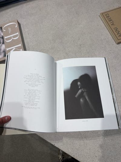 The image shows an open book lying on a light grey surface. The book contains a black and white photograph on the top page, labeled "Nicole 1985." The bottom page contains poetry. Two other books are partially visible near the primary book: one labeled "ANNI ALBERS" and another with "C-BK SUNITA KUMAR NAIR" visible on its cover. A finger is visible in the lower right corner of the frame. The location is not specified in the image.