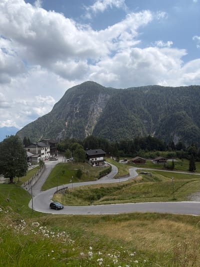 The image shows a panoramic view of a mountain village, likely in Sauris / Zahre, Italy, under a cloudy sky. In the foreground, a winding road curves through a grassy hillside dotted with wildflowers. A dark-colored car is driving on the road. In the mid-ground, several buildings with traditional alpine architecture are visible, including a multi-story white building with brown trim and a darker building with a pitched roof. Trees and vegetation surround the buildings. The background is dominated by a large, steep mountain covered in dense evergreen forest, with patches of exposed rock face. The overall scene depicts a rural mountain landscape with a small settlement.