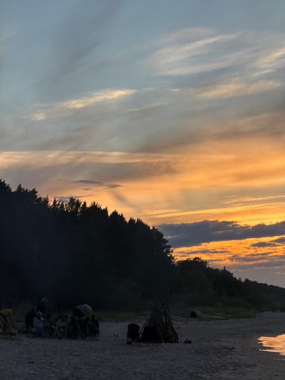 The image depicts a serene sunset scene at Plieņciems, Latvia. The sky is ablaze with hues of orange and gold, with streaks of light illuminating the wispy clouds. The sun, though not directly visible, casts a warm glow across the horizon, indicating the late afternoon or early evening.

In the foreground, a group of people are gathered on a sandy beach, seated in camping chairs. They appear to be enjoying a quiet evening, perhaps a bonfire or a casual get-together, as evidenced by the presence of what looks like a fire pit and some bundled wood. The silhouettes of trees form a dark, lush backdrop against the vibrant sky, suggesting a coastal forest adjacent to the beach.

The calm atmosphere and the warm, diffused light evoke a sense of peace and relaxation. The gentle lapping of water might be implied by the reflective quality of the surface on the right side of the image, suggesting proximity to the sea. No visible text is present in the image.