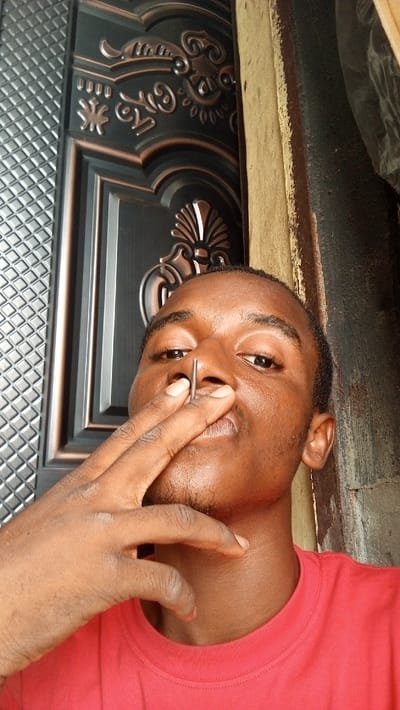 A male individual with dark skin, wearing a red t-shirt, is depicted from the chest up, facing the camera. His left hand is raised, with his thumb and index finger holding a slender, silver-colored metal pin vertically at his nose. The tip of the pin is positioned near or against his right nostril. The background features a dark-colored, ornate metal door with copper or bronze-toned embossed patterns, including a diamond-textured panel on the left and various scrollwork designs on the right. To the immediate right of the door and behind the individual, a light brown wooden structure, possibly a door frame or wall section, is visible. The scene appears to be an indoor or sheltered outdoor setting in Ogbunike, Nigeria.