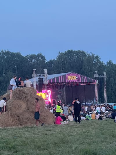 This is an outdoor event taking place in Surakovo, Russia, with a stage set up in the background. In the foreground, a large pile of hay bales serves as a seating area for some of the attendees. Several people are visible on and around the hay bales, some are sitting, some are standing, and others are climbing on them. In front of the hay bales, more people are gathered, sitting and standing on a grassy field. The stage has a pink and black striped canopy with the word "ПЛЯЖ" (Plage) written in large, yellow letters. Pink and purple lights are illuminating the stage. Trees form a backdrop behind the stage and the event area. The sky is a clear, muted blue, indicating it is either early morning or late evening. The overall scene depicts a lively outdoor festival or concert.