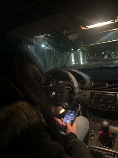 The image depicts the interior of a stationary BMW vehicle at night, located in the Glyka Nera Municipal Unit, Greece. A person with long dark hair, wearing a dark jacket with a fur-trimmed collar, is positioned in the driver's seat. Their right hand is engaged with a dark-colored smartphone, which displays an illuminated screen with multiple colorful application icons. The vehicle's dashboard includes a BMW-branded steering wheel fitted with a carbon fiber-style cover, an instrument cluster, and a center console featuring an integrated radio/CD player and manual gear shifter, the latter adorned with a red and white checkered fabric accessory at its base. An overhead interior light is active, illuminating parts of the cabin. Through the windshield, a nighttime street scene is visible, illuminated by artificial streetlights, revealing trees and solid structures lining the road, consistent with an urban or suburban environment.