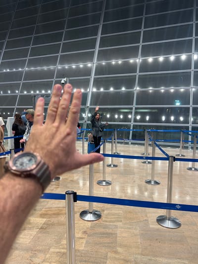 The image depicts an interior view of an airport terminal in Israel, specifically identified by Hebrew text visible on the blue queue barriers. In the immediate foreground, a male's blurred left hand is extended towards the camera, revealing a rose gold-toned digital/analog wristwatch on the wrist.
In the midground, a young woman stands center-right, smiling and waving her raised right hand. She is wearing a dark jacket, dark trousers, and a black shoulder bag, with a light blue rolling suitcase positioned at her left side. To the left of the scene, behind the foreground hand, several other individuals are partially visible in a queue.
The environment is structured by a series of silver stanchions with retractable blue fabric belts, forming multiple queuing lanes. One stanchion on the far right has a small "SECURITY" label. The floor is tiled with large, rectangular light-brown and greyish patterns. The background features a vast, multi-paned glass curtain wall, reflecting interior lights and allowing a glimpse of faint exterior lights, suggesting an evening or nighttime setting. A dome-shaped security camera is mounted on a pole against the glass wall in the background.