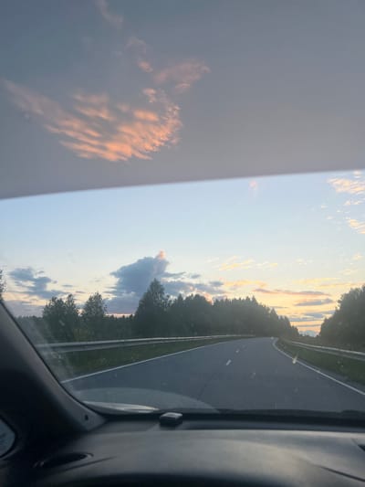 The image depicts a view from inside a vehicle, looking out at a highway winding through a forested landscape at dusk. The sky above is a soft gradient of blues and grays, with wisps of pink and orange clouds, suggesting the sun is setting. The road itself is paved, with white lane markings, and curves to the right. On either side of the road are guardrails and dense trees. The interior of the car, including the dashboard and windshield frame, is visible in the foreground, indicating the perspective of the photographer. The overall scene is serene and natural, characteristic of a rural or semi-rural setting. The location is identified as Kliuchevka 2-Ya, Russia.