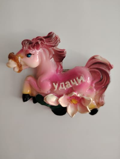 A pink, glossy decorative figurine of a horse is depicted against a plain light background. The horse is in a dynamic, rearing pose, with its front legs lifted. It features blue eyes and a darker pink/magenta mane and tail. At the base of the figurine, there are two light-colored flowers with yellow centers and some green leaves. The word "УДАЧИ" (UDACI), which translates to "Good Luck" in Russian, is inscribed in raised white capital letters on the side of the horse's body. The object appears to be a magnet or small ornament, possibly made of ceramic or resin, and shows some minor signs of wear and dust. The location associated with this media is Сусады-Эбалак, Russia.
