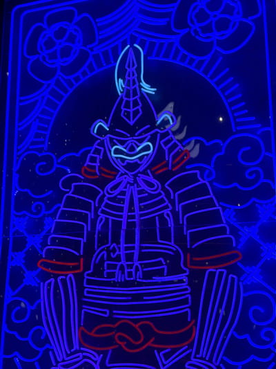 The image displays a vibrant blue neon light installation depicting a stylized samurai. The samurai is rendered in outline form with a menacing expression, wearing traditional armor. Red neon accents highlight parts of the armor, such as the wristbands and a sash. The background features a dark, starry sky with stylized clouds and floral motifs, also outlined in blue neon. The composition is symmetrical and centered on the samurai figure. The image was taken at night on Carretera De San Martín, Spain, where this neon art is likely a part of a festive decoration or public art display. There are no people or active interactions visible in the scene.