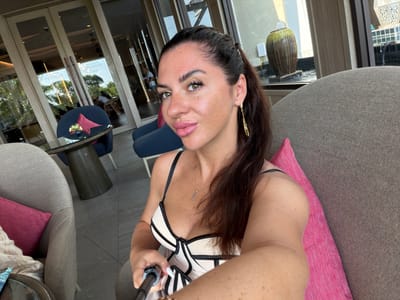 A woman with long, dark hair pulled into a ponytail, wearing a black and white corset-style top and gold earrings, is taking a selfie in what appears to be a hotel lobby or lounge area in Bang Muang, Thailand. She is sitting in a plush, grey armchair with a pink cushion. In the background, through glass doors, trees and other patrons are visible. A decorative urn and fountain are also present in the outdoor area. The woman is holding a selfie stick, and her expression is relaxed. The lighting suggests it is daytime, and the overall ambiance is one of leisure.