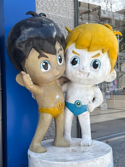 Two cartoon-like statues of children are depicted, appearing to lie side-by-side. The image is oriented vertically, so the figures are arranged one above the other in the frame. The upper statue has bright yellow hair and light-colored skin, with large blue eyes and a cheerful smile. It wears blue shorts featuring a green circular emblem. Its body shows noticeable cracks and wear.
The lower statue has dark hair and tan skin, with similar large blue eyes and a gentle smile. It wears yellow shorts adorned with a red circular emblem. This statue also exhibits significant surface cracks and deterioration. Both figures have their arms bent, with the upper figure's right hand (as viewed from the top of the statue) resting against a white, cylindrical base or pillar that supports both figures. The lower figure's feet also rest against this cylindrical base.
The background shows reflections of modern architectural elements, primarily glass windows of buildings, indicating an urban outdoor setting, possibly a commercial area. The lighting suggests it is daytime. The statues appear to be part of a public art installation or display, possibly mascots, and their weathered appearance indicates exposure to the elements over time. No legible text is visible on the statues themselves or their immediate bases. The overall scene conveys a sense of whimsical public decoration.