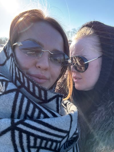 Two individuals are depicted in a close-up outdoor portrait, likely in Mysiadło, Poland. The prominent figure, positioned to the left, is an individual with dark hair, wearing rimless, dark-tinted sunglasses with gold-colored frames featuring an animal head motif on the temples; a reflection on the lens shows the text "LOSTERM". This person is wrapped in a light gray and black geometrically patterned blanket or scarf. Partially visible to the right, and slightly behind, is a second individual wearing dark, oval-framed sunglasses with thick black rims. This second person's face is partially obscured, and their mouth is slightly open, suggesting an interaction. They are wearing a dark, possibly hooded, textured outer garment. The scene is illuminated by strong, direct sunlight from the upper left, causing lens flare and highlighting the subjects' hair. The background consists of a clear blue sky and indistinct, bare trees, implying an outdoor setting.