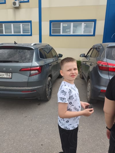 The image shows a young boy standing in front of a building with cars parked nearby. He is wearing a white t-shirt with a black graphic print and black pants. The boy is holding a smartphone in his right hand and looking towards the camera with a slight smile. The background consists of a building with yellow walls and blue accents, and two dark-colored SUVs. One SUV, a dark grey Skoda, is parked to the left of the boy, and another dark SUV is to his right. The ground is asphalt. The lighting suggests it is daytime, possibly overcast. The building has several windows with blue frames. There is text visible on the rear of the Skoda, including "SKODA", "HY 152", and "RUS". There is also "4X4" printed on the lower right of the Skoda's rear bumper. The t-shirt the boy is wearing has repeated text and graphics, including the word "SUPER". The overall scene appears to be an outdoor setting in a parking area.