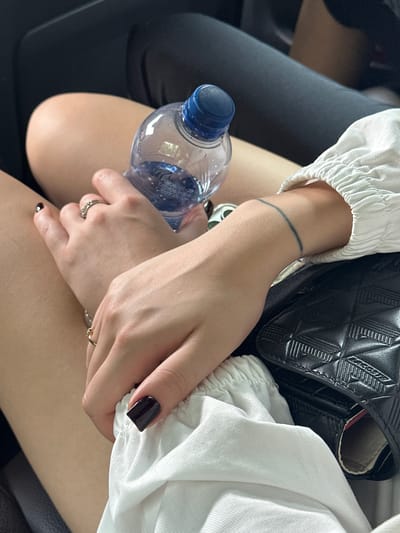 The image shows a close-up view of two women's arms and legs. One woman has dark nail polish and is wearing a white ruffled shirt. She is holding a bottle of water and has rings on her fingers. The other woman has a thin black line tattoo on her forearm. They appear to be sitting together, possibly in a vehicle, given the dark background that resembles upholstery. The overall lighting suggests an indoor or shaded outdoor setting. There is no specific indication of the location being in Qingcheng District, China.
