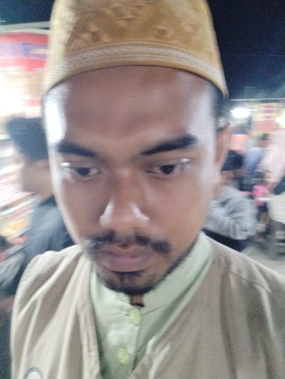 This media file is a close-up, slightly blurry photograph of a young man wearing a traditional Muslim kufi hat and a light-colored shirt. The man has dark hair, a prominent mustache, and a beard. His eyes are looking downwards. The background is out of focus, showing what appears to be a busy street scene with blurred lights and people. The overall impression is that this is a candid, informal photograph taken outdoors, possibly at night given the lighting.
The subject's attire suggests he may be participating in or attending a religious event or simply wearing traditional clothing. The blurred background indicates movement, suggesting the photo was taken while walking or in a dynamic environment. There are no discernible landmarks or specific location cues visible in the image, aside from the general context of a populated area. The lighting suggests it could be evening or night. No text is visible in the image.