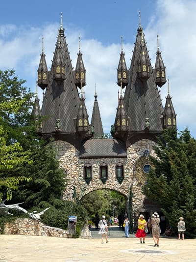 The image shows the entrance gate of the Castle Ravadinovo in Ravadinovo, Bulgaria, on a sunny day. The gate is a grand structure with multiple conical towers topped with spires and adorned with decorative elements. The facade is constructed from rough-hewn stone blocks and features arched doorways and windows. Lush green trees and foliage surround the castle, framing the entrance. Several people are visible in the foreground and middle ground, some walking, others standing, dressed in casual summer attire. One person in a red skirt and yellow top is looking towards the camera. A large, stylized dragon sculpture is present on the left side of the image. The sky is blue with scattered clouds.