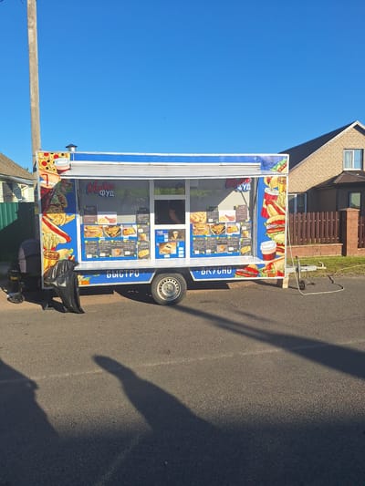 An outdoor, eye-level, full shot shows a blue and white food truck parked on an asphalt road during the day. The food truck is adorned with colorful graphics of food items like pizza, hot dogs, and fries, and features a menu displayed on its side. The words "Make фуд" and "Быстро" are visible on the truck. A person is partially visible inside the truck, likely serving customers. To the left of the truck, a black trash bag lies on the ground next to a small cart with wheels. In the background, a brick house with a brown fence is visible, and a wooden utility pole stands to the left. The sky is clear and blue. The location is Цімкавіцкі сельскі Савет, Belarus.