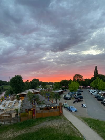 This is a high-angle, wide shot of the "BODEGA BEACH CLUB" at sunset in Rinteln, Germany. The sky is filled with dramatic, colorful clouds in shades of grey, pink, and orange. In the foreground, the beach club features thatched roofs, palm trees, and lounge chairs. Adjacent to the club is a parking lot filled with various cars and a few motorcycles. A river or body of water is visible to the left of the beach club, with trees lining its bank. A paved pathway leads from the foreground grassy area towards the beach club. The overall scene depicts an outdoor recreational venue during a colorful sunset.
