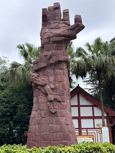 This forensic image captures a monumental, reddish-brown stone sculpture situated in a landscaped park environment in Qingyang District, China, under an overcast sky. The sculpture, crafted from rough-hewn, irregular blocks of stone, occupies the central and left portions of the frame, towering from the bottom edge almost to the top. Its form suggests an abstract, upward-reaching structure, with the uppermost section terminating in multiple, stylized, finger-like projections. The surface of the stone is rugged and textured.

Behind and to the right of the sculpture, a prominent array of palm trees with long, drooping fronds is visible. Further back and to the left, dense green foliage comprising various trees and bushes fills the background. A traditional-style building is partially discernible in the background to the right, featuring white walls accented by dark red cross-beams and a dark, gable roof.

In the lower right quadrant, positioned behind a neatly trimmed row of light green bushes that forms the immediate foreground, two rectangular signs with Chinese script are visible. The more prominent sign on the right has an ornate, reddish-brown frame with a spiral motif and larger characters that translate to "Inherit the red gene, cherish the revolutionary martyrs," indicating a memorial or historical context. The ground at the base of the sculpture and in the foreground is covered with a uniform layer of vibrant green low-lying bushes. No individuals are present in the scene.