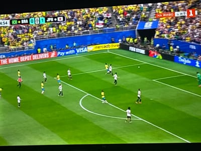 This is a still image captured from a television screen displaying a football (soccer) match. The scene is a brightly lit football stadium, with a vibrant green pitch and a large crowd of spectators in the background, many wearing yellow and green attire.
The primary focus is the football match in progress. Several players, dressed in either yellow and blue kits or white and black kits, are visible on the field, engaged in the game. One player in a yellow kit is near the penalty box, while others are spread across the pitch, in various positions and actions.
At the top of the screen, a scoreboard graphic indicates the match is between "BRA" (Brazil) and "JPN" (Japan), with the score currently at 0-1 in favor of Japan. A timer shows 4:54. Below the scoreboard, advertisements are visible along the sidelines, including "THE HOME DEPOT DELIVERS" and "Pay with VISA." Further along, "mercado livre" and "FIFA STORE" are also advertised.
To the right of the screen, there's a red banner with white Cyrillic text that reads "НА ЖИВО," meaning "Live" in Russian. Adjacent to this, a smaller graphic displays "5.H.T 1," potentially indicating a broadcast time or score detail. The overall atmosphere appears to be energetic, typical of a live sporting event. The image seems to be taken from a slightly elevated angle, looking down onto the pitch. No specific weather conditions are apparent.