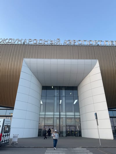 This image shows the entrance of what appears to be the Platov International Airport in Kitsjanovo, Russia, based on the Cyrillic text visible on the building's facade. The architectural style of the entrance includes a modern design with a combination of curved wooden panels and rectangular white panels. A glass facade is centered in the entrance. Several individuals are walking into or out of the airport. A white van with distinctive black and white patterned graphics is parked near the entrance with a luggage cart next to it. The sky is clear and blue.
