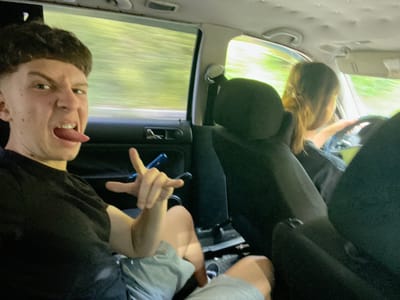 This is a candid, medium-shot, eye-level photograph taken from inside a moving vehicle. The perspective is looking towards the front of the car. In the foreground, a young Caucasian male with short, styled dark hair is making a face at the camera, sticking out his tongue which has braces. He is wearing a black t-shirt and grey shorts, and is positioned on the left side of the frame. His right hand is raised, with two fingers extended in a peace sign, and he is holding a blue object. Behind him, to the right and slightly blurred, is a young Caucasian female with long, light brown hair, who appears to be driving. She is wearing a black top and is holding the steering wheel. The interior of the car is visible, including dark upholstery and the car's ceiling. The windows show a blurred background of green foliage, suggesting the vehicle is traveling on a road surrounded by trees. The lighting is natural and bright, indicative of daytime. There is no discernible information to place this image specifically within the city of Vlahovo, Bulgaria, or any other specific urban location.