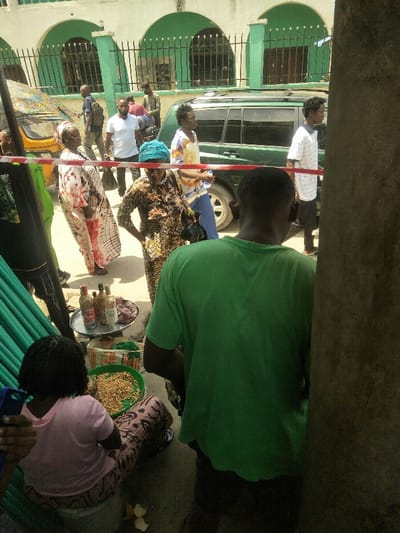 The image depicts an outdoor street or market scene in Ajah, Nigeria, under bright daylight.
In the immediate foreground, to the left, a person with dark braided hair, wearing a pink top and patterned brown skirt, is seated on a low object, facing away from the camera. A blue smartphone is partially visible in their left hand. On the ground next to this individual, a green plastic basin filled with light-colored, elongated edibles (consistent with groundnuts or peanuts) is present, alongside a woven basket. A metal tray holds five glass bottles containing various liquids and a pile of reddish-brown material. To the right, the back of a male subject in a bright green t-shirt is prominently visible, standing near a dark concrete pillar that frames the right side of the image.
The midground features an unpaved road or thoroughfare where multiple individuals are present and actively moving. A red and white barrier tape extends horizontally across this area. Pedestrians include a woman in a white and red patterned dress, a woman in a dark patterned dress with a teal headscarf holding a black bag, and several men in casual attire, one in a white t-shirt and another in a white striped t-shirt. A dark green SUV is positioned behind the barrier tape on the right side of the road, and part of a yellow Keke Napep (motorized tricycle) is visible on the far left behind the barrier.
The background reveals a light-colored building with distinctive green architectural elements, specifically multiple arched structures above window or door openings, which are secured with dark metal grilles.