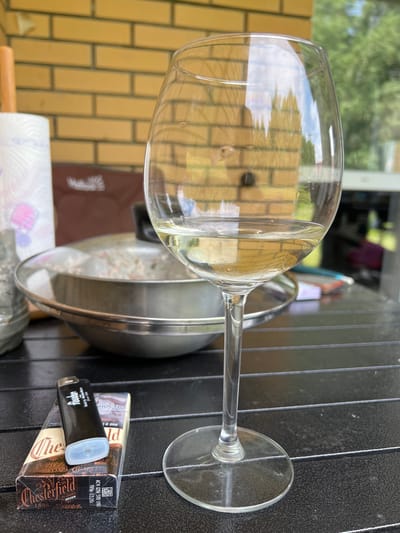 A glass of white wine is pictured on a dark, textured table, with a pack of Chesterfield cigarettes and a black lighter in the foreground. A silver bowl with a lid, seemingly containing food, is behind the wine glass. In the background, a brick wall and lush green trees are visible. The scene appears to be an outdoor setting, possibly a patio or balcony. The lighting suggests it is daytime. The image was captured in Novosibirsk, Russia.