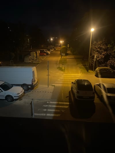 The image shows a street scene at night in Konstantynów Łódzki, Poland. A row of parked cars is visible on the left side of the road, including a white van and a white passenger car. A pedestrian crossing with white stripes is illuminated by streetlights. On the right side of the road, there are more parked cars, including a dark-colored SUV and two lighter-colored passenger cars. Trees line the street, and there are streetlights casting a yellow glow on the scene. The sky is dark, indicating it is nighttime.