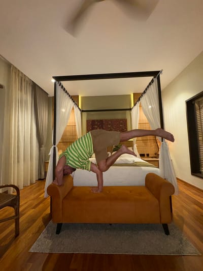 An adult male is performing a balancing pose indoors within what appears to be a hotel room, consistent with boutique accommodations found in George Town, Malaysia. The man, barefoot and wearing a green and white striped short-sleeved shirt with khaki shorts, is inverted. His head is resting on his forearms, which are placed on the left end of a rectangular orange upholstered bench. His right hand is flat on the bench's surface. His left leg is bent at the knee, with the foot near his hip, while his right leg is fully extended horizontally backward, suspended in the air.
The room features a large canopy bed with white drapes, positioned centrally behind the man. The bed has a white comforter and a green folded blanket or runner at its foot. Above the bed's headboard, a red and gold patterned textile art piece is displayed, flanked by bamboo blinds and recessed wall lighting. To the left, light-colored sheer curtains cover a window or door. To the right, a window with dark blinds is visible. The floor is polished wood, and a light grey rectangular rug is positioned beneath the orange bench. A ceiling fan, blurred by motion, is visible overhead. The lighting is warm and appears to be artificial. A partial view of a wicker chair is visible in the lower-left corner of the frame.