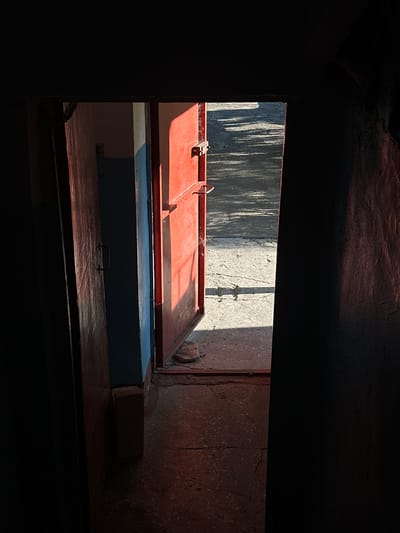 The image depicts an open doorway leading to an outdoor area. The doorway is framed by dark, textured walls on the sides, suggesting a building interior. A red metal door is ajar, revealing a sunlit concrete path outside with shadows cast by an unseen object. Inside the doorway, a wooden door is also partially open, with a dark handle visible. A blue-painted wall segment and a brown brick are present within the interior space near the doorway. The lighting suggests late afternoon or early morning sun. The overall impression is of a humble, functional entrance in a city environment. Based on the context of Podilsk, Ukraine, this could be an entrance to a residential building, a utility area, or a commercial space.