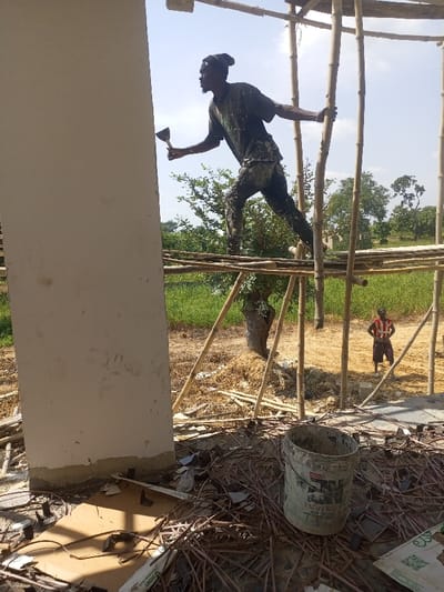 The scene depicts an outdoor construction site in Gidan Kado, Nigeria, under a clear, bright sky. In the midground, a dark-skinned male individual, dressed in a dark beanie, a paint-splattered dark t-shirt, and dark trousers, stands on a bamboo scaffold. He is captured in profile, facing left, holding a small, flat metallic tool (possibly a scraper or trowel) in his right hand while his left hand grips a vertical bamboo pole for support, suggesting movement or balance. To his left, a large, light-colored concrete column or wall of a building under construction dominates the left side of the frame.
Behind the scaffold, green foliage, including trees and dense vegetation, forms a natural backdrop, with one prominent gnarled tree trunk visible centrally. Further back and to the right, a second dark-skinned male figure stands on the dry, light-brown ground. He wears a red and white striped shirt and dark shorts, with his hands positioned on his hips, observing the scene to his left.
The foreground is littered with construction debris, including pieces of wood, cardboard, metal rebar, and other discarded materials. A dirty, white cylindrical bucket, marked with "EMULSION," sits prominently among the debris on the ground level, near the base of the concrete column. The overall environment suggests ongoing manual construction work in a rural or semi-urban setting.