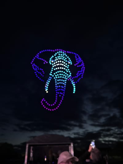 A nighttime image shows a lit-up drone display in the shape of a jellyfish or similar marine creature. The drones emit a blue, white, and purple light and are arranged in a pattern to define the shape. The jellyfish shape appears to be centered and positioned above the people watching. The location appears to be somewhere outdoors where people are watching the light display. The city is South Norfolk, United Kingdom. Some people are visible on the right side of the image, including one individual holding up a phone, possibly to record or photograph the drone display. The backdrop is a dark sky with some cloud cover or structures visible on the lower right.