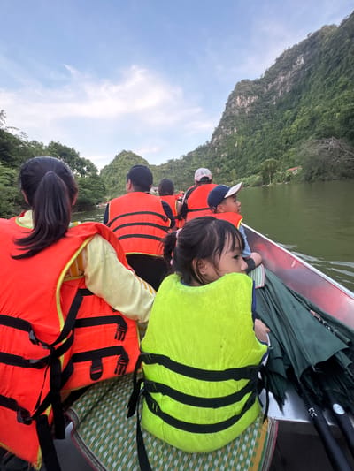 A boat filled with people, including children and adults, travels down a river with lush green mountains on either side. The people are wearing orange and green life vests for safety. The sky is cloudy with hints of blue. The boat appears to be in motion, as indicated by the water's reflection. This scene likely takes place in the karst landscape of Yên Sơn Ward, Vietnam, known for its scenic rivers and mountains.