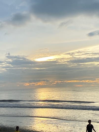 The image shows a beach scene at Bateetimoh, Indonesia. The sky is filled with dramatic clouds, with a bright band of sunlight breaking through them, casting a golden reflection on the ocean. Gentle waves are washing onto the sandy shore. In the foreground, two young individuals are visible on the beach. One, wearing a yellow shirt, is partially obscured. The other, wearing a dark shirt, is walking towards the right side of the frame. The overall atmosphere is serene, likely during sunrise or sunset.