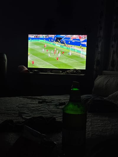 The image depicts a dimly lit bedroom in the city of Karadjovo, Bulgaria. A television screen in the background displays a soccer match, with players in red and white uniforms on a green field. The score is visible as "0-0." In the foreground, a green glass bottle, presumably containing a beverage, stands upright on a bed. The bed is covered with dark-colored sheets and a lighter-colored patterned duvet. A dark object, possibly a remote control, lies on the bed near the bottle. A pink baseball cap is placed on a shelf beneath the television, alongside other indistinct objects. The room's ambiance is dark, with the only significant illumination emanating from the television screen.
